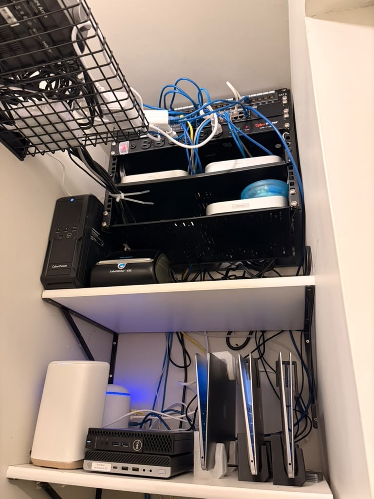 Spicer's server closet: a small network rack on the top shelf, and used MacBooks standing upright in 3D printed stands on the bottom shelf